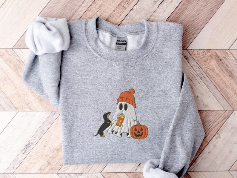 May include: A gray sweatshirt with a cute ghost wearing a red hat and holding a cup of popcorn. A small black dog is standing next to the ghost. A pumpkin is in front of the ghost.
