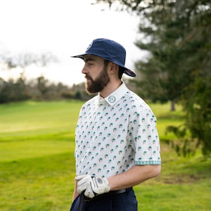 May include: A man wearing a white and green patterned short-sleeved shirt with a blue bucket hat, a white golf glove, and navy blue trousers. He is holding a golf club.