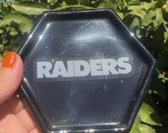 Puede incluir: Una bandeja hexagonal negra con la palabra "RAIDERS" en letras blancas. La bandeja se sostiene contra un fondo de hojas verdes y luz solar.
