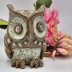 May include: A ceramic owl figurine featuring a glossy brown glaze with cream-colored accents around the large circular eyes and textured belly. The owl has prominent ear tufts, a row of small raised dots across the chest, and detailed feet.