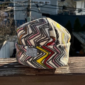 May include: A folded beanie hat with a colorful zigzag pattern in shades of gray, pink, yellow, and red. The hat has a small rectangular tag with the text "AFTER MOON". The hat is made of a soft, textured material.