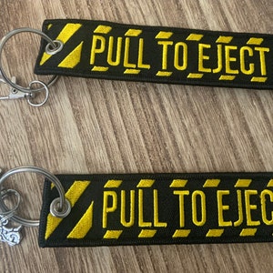 May include: Two rectangular black fabric keychains with yellow embroidered borders and the text PULL TO EJECT in yellow. Each keychain features a silver metal split ring and a lobster clasp. One keychain includes a small silver motorcycle charm.