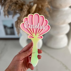 Könnte beinhalten: Ein rosa-weißes Namensschild aus Acryl in Muschelform mit dem Namen "Caroline" in weißer Schrift. Der Griff ist perlmuttgrün. Der Hintergrund ist verschwommen, mit neutralen Tönen.