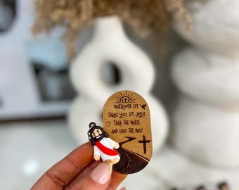 May include: A wooden oval keychain with a brown background and a white figure of Jesus walking on a road. The keychain has the text "WHEREVER LIFE TAKES YOU, LET JESUS TAKE THE WHEEL AND LEAD THE WAY." with a cross at the bottom.