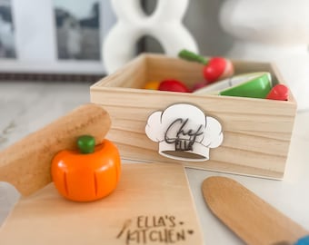 May include: Wooden toy kitchen set with a chef's hat logo, a wooden box filled with toy vegetables, a cutting board with "Ella's Kitchen" text, a toy pumpkin, and a wooden knife. The set is designed for children's play.