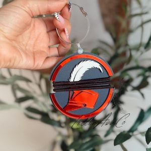 May include: A circular ornament featuring a dark gray base with a metallic orange border and a central cutout design of a helmet with a white feather plume. The piece is wrapped with two dark brown elastic bands and hangs from a silver wire loop.
