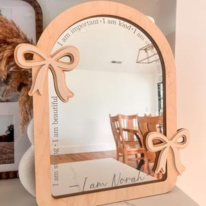 May include: Wooden framed mirror with an arched top and two bow accents. The mirror has the text "I am important, I am kind, I am smart, I am strong, I am beautiful, I am brave, I am loved" and "- I am Norah" etched on the surface.