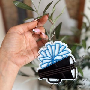 May include: A white acrylic keychain featuring a blue cheerleading pom-pom graphic layered over a black megaphone silhouette. The charm is attached to a silver-toned metal cable ring. The text Little Ash Co is printed in black script at the bottom.