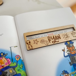 May include: A wooden bookmark with the name "RYAN" and the phrase "This butt on the loose will never do!" is placed on an open book. The bookmark has engraved alien, star, game controller, sun, and monster icons.