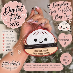 Puede incluir: Una etiqueta para bolso en forma de dumpling que sostiene una coleta, con una parte superior blanca con cara sonriente, una base color canela con el texto 'THROW YOUR HAIR UP. WE'RE SQUISHY HUNTING.' y una goma negra, sujeta a una anilla metálica.