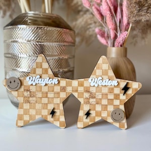 May include: Two wooden star-shaped baby announcement signs. Each star has a checkered pattern and includes the names "Waylon" and "Weston" in white script. The signs have spaces for date, time, weight, and length, with smiley face and lightning bolt accents.