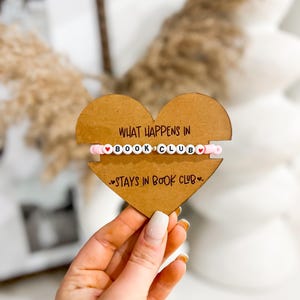 Puede incluir: Un letrero de madera en forma de corazón con el texto "What happens in book club stays in book club" y una pulsera de cuentas rosadas con cuentas de letras blancas que deletrean "book club" y cuentas de corazón rojas.