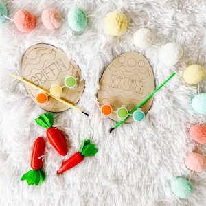 May include: Two wooden Easter egg shaped paint palettes with the names Griffin and Holden written on them. The palettes are surrounded by colorful pom poms and paint pots.
