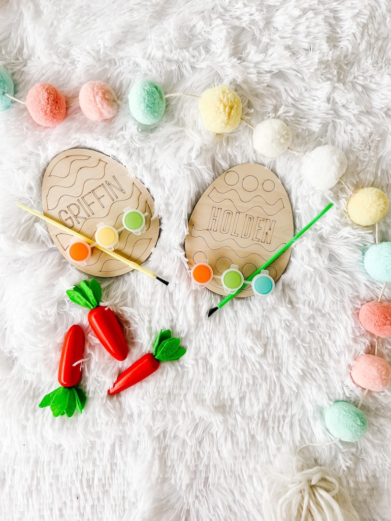 May include: Two wooden Easter egg shaped paint palettes with the names Griffin and Holden written on them. The palettes are surrounded by colorful pom poms and paint pots.