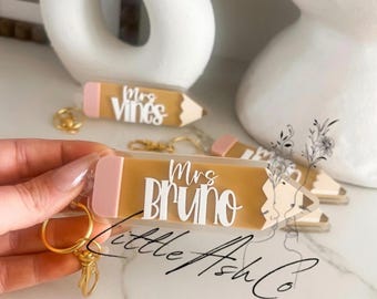 May include: A pencil-shaped acrylic keychain featuring a gold-colored body, a pink eraser end, and a white pointed tip. The keychain displays the white text 'Mrs Bruno' in a script font and includes a gold-toned metal split ring and lobster clasp.
