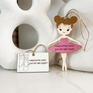 May include: Wooden ballerina ornament with a pink tutu and brown hair. The ornament has the text "Congratulations, Dancer - Your light shined bright!" on the front.