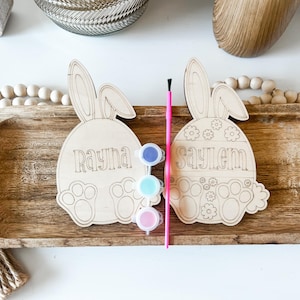 May include: Two unfinished wooden bunny-shaped Easter egg ornaments with the names Rayna and Saylem written on them. The ornaments are on a wooden tray with paint pots and a paintbrush.