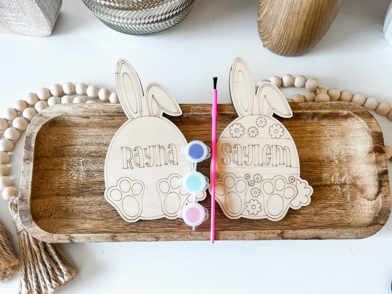 May include: Two unfinished wooden bunny-shaped Easter egg ornaments with the names Rayna and Saylem written on them. The ornaments are on a wooden tray with paint pots and a paintbrush.
