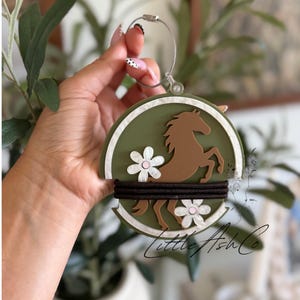 May include: A circular olive green ornament featuring a brown horse silhouette, two white flowers with pink centers, and a white border. A black elastic hair tie is wrapped horizontally across the center. The ornament hangs from a silver wire loop.