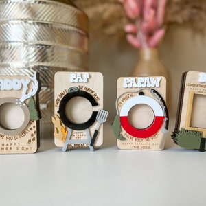 May include: Four wooden photo frames with different designs and text. The frames are decorated with images of deer antlers, a grill, a fishing bobber, and a football goal post. The text on the frames reads "Daddy", "Pap", "Papaw", and "Dad".