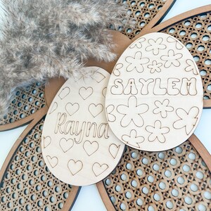 May include: Two wooden Easter egg ornaments with cut-out designs. One egg has hearts and the name "Rayna" and the other has flowers and the name "Saylem".