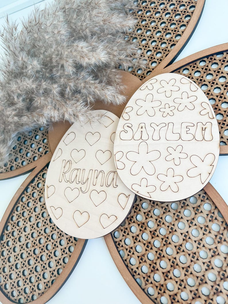 May include: Two wooden Easter egg ornaments with cut-out designs. One egg has hearts and the name "Rayna" and the other has flowers and the name "Saylem".