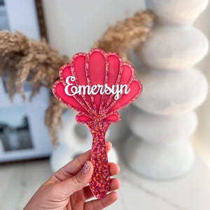 May include: A pink seashell-shaped hairbrush with the name "Emersyn" in white script. The brush handle and shell are covered in glitter. The brush is held up against a blurred background.