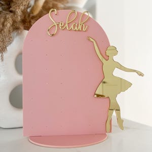 May include: A pink earring display stand with a gold ballerina silhouette and the name "Selah" in gold script. The stand has a rounded arch shape with small holes for earrings and a circular base. The background is a soft white.