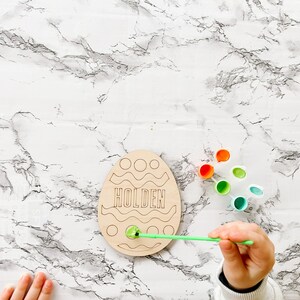 May include: A wooden Easter egg shaped craft with the name Holden written on it. The egg is being painted with green paint using a paintbrush. There are several small containers of paint in different colors.