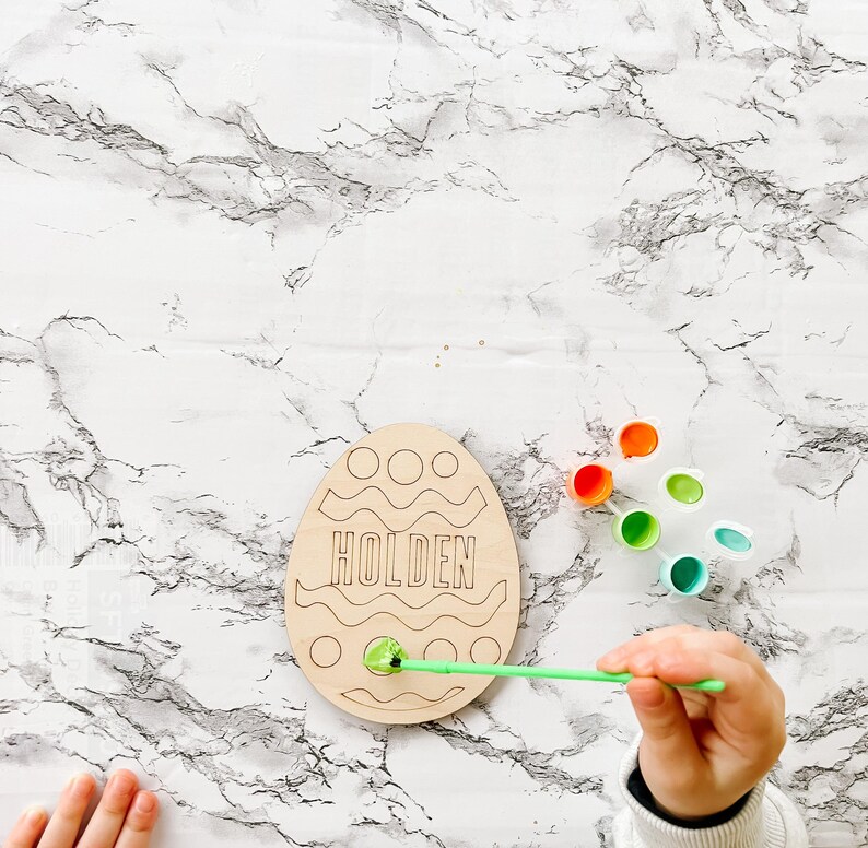 May include: A wooden Easter egg shaped craft with the name Holden written on it. The egg is being painted with green paint using a paintbrush. There are several small containers of paint in different colors.