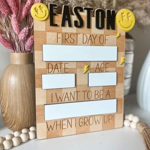 May include: Wooden sign with the name "EASTON" in black letters, yellow smiley face accents, and space to write "FIRST DAY OF", "DATE", "AGE", and "I WANT TO BE A WHEN I GROW UP".