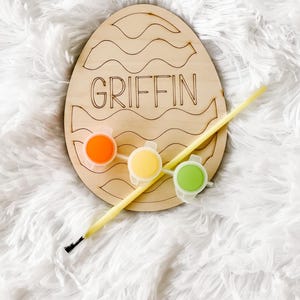 May include: Wooden Easter egg craft kit with the name "GRIFFIN" painted on it. The kit includes three small paint pots in orange, yellow, and green, and a paintbrush.