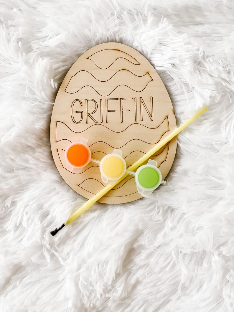 May include: Wooden Easter egg craft kit with the name "GRIFFIN" painted on it. The kit includes three small paint pots in orange, yellow, and green, and a paintbrush.