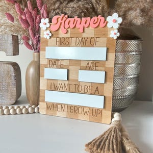 Könnte beinhalten: Ein Holzschild mit dem Namen "Harper" in Rosa und Weiß, mit Blumenakzenten. Das Schild hat Felder für "First Day Of", "Date", "Age" und "I Want To Be A When I Grow Up!" in einem Schachbrettmuster.