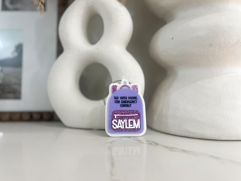 May include: A purple backpack-shaped tag with glitter accents and the name "SAYLEM" in white. The tag includes the text "TAP WITH PHONE FOR EMERGENCY CONTACT".