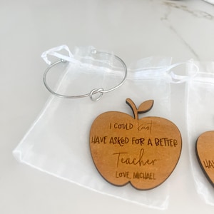 May include: A wooden apple-shaped ornament with the text "I could not have asked for a better teacher. Love, Michael" engraved on it. The ornament is in a white organza bag with a silver knot bracelet.