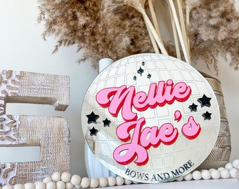 Pode incluir: Um espelho de bola de discoteca prateado com uma inscrição "Nelle Jae's" em rosa e vermelho e estrelas pretas. O espelho tem um padrão de grade e o texto "Bows and More" está impresso na parte inferior.