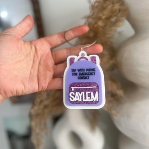 May include: A purple backpack-shaped tag with white trim and the name "SAYLEM" in white letters. The tag has a silver loop and the text "TAP WITH PHONE FOR EMERGENCY CONTACT".
