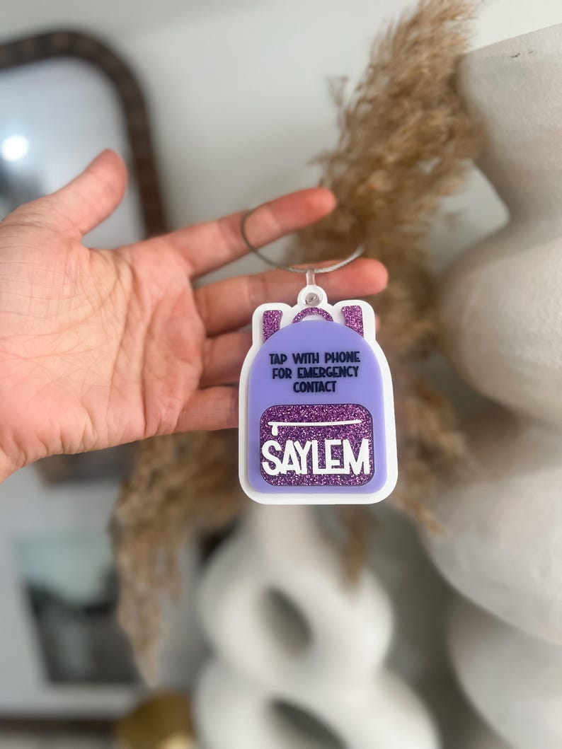 May include: A purple backpack-shaped tag with white trim and the name "SAYLEM" in white letters. The tag has a silver loop and the text "TAP WITH PHONE FOR EMERGENCY CONTACT".