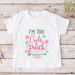 May include: White t-shirt with a pink and green graphic that says "I'm Too Cute To Pinch". The graphic includes shamrocks and an arrow.