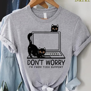 May include: A heather gray t-shirt featuring a graphic of a laptop with two black cats. The text on the shirt reads "DON'T WORRY I'M FROM TECH SUPPORT". The shirt is hanging on a wooden hanger.