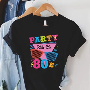 K&ouml;nnte beinhalten: Schwarzes T-Shirt mit einem bunten Grafikdesign, auf dem "Party Like The 80's" mit einer Sonnenbrille in der Mitte des Textes steht.