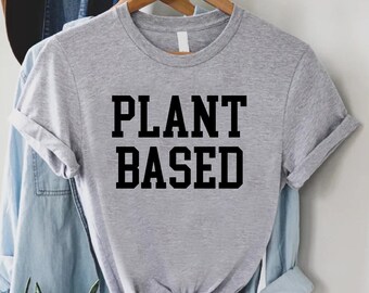 Puede incluir: Una camiseta gris con el texto "PLANT BASED" impreso en negro.