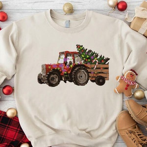 May include: A cream colored sweatshirt with a red tractor pulling a Christmas tree. The tractor is decorated with purple, green, and red ornaments. The tree is decorated with red and green ornaments.