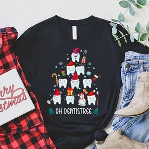 以下が含まれることがあります： 歯でできたクリスマスツリーのデザインが施された黒いTシャツ。ツリーはオーナメント、キャンディーケーン、雪だるまなどで飾られています。ツリーの下には「Oh Dentistree」という文字が印刷されています。