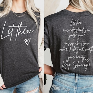 May include: A dark gray t-shirt with white text that reads "Let them" on the front and "Let them misunderstand you judge you gossip about you never doubt your worth your beauty Keep Shining!" on the back. The shirt has a small heart symbol next to the text on the front and back.