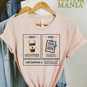 May include: A pink t-shirt with a graphic design that says "First Coffee, Then Data, I am earning a break". The graphic features a cup of coffee and a clipboard with a pen.