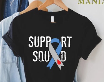 Puede incluir: Camiseta negra con una cinta azul y gris con una gota de sangre roja. El texto "SUPPORT SQUAD" está impreso en blanco en la camiseta.