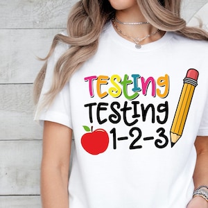 May include: White t-shirt with the text "Testing Testing 1-2-3" in rainbow colors. A red apple and a yellow pencil are next to the text.