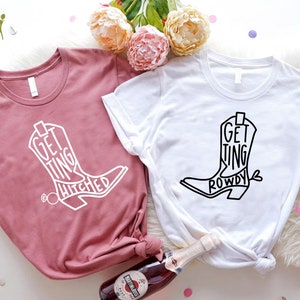 May include: Two t-shirts with a cowboy boot design. One shirt is pink with white text that says "Getting Hitched". The other shirt is white with black text that says "Getting Rowdy".
